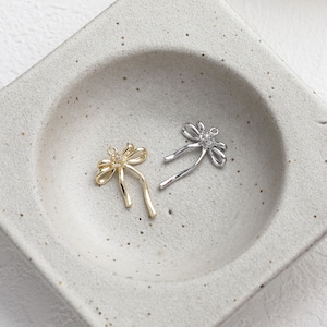 May include: Two bow-shaped charms, one in a polished gold tone and one in a polished silver tone. Each charm features a small, clear, circular stone set at the centre of the bow and a small metal loop at the top for attachment to jewellery.
