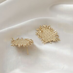 May include: A pair of gold-toned earrings with a woven, textured design. The earrings have a frayed, irregular edge.