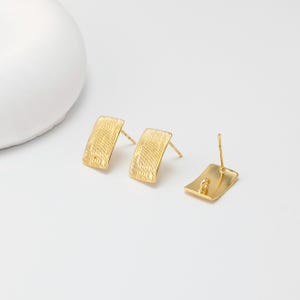 May include: Gold-coloured rectangular stud earrings with a textured surface. The earrings are displayed on a white background. The earrings have a post and butterfly backing. The earrings are a simple, yet elegant design.