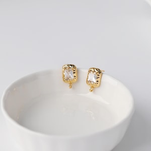 May include: A pair of gold-toned stud earrings featuring a rectangular, clear faceted gemstone set within a textured, hammered metal frame. Each earring includes a small circular loop at the bottom, designed for attaching additional jewelry components.