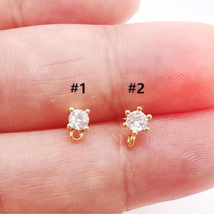 May include: Two small gold-colored jewelry findings with clear, round gemstones. One has a square setting, the other a star-like setting. Both have a small loop at the bottom for attaching to other jewelry components.