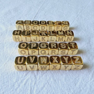 Könnte beinhalten: Ein Satz von 26 goldfarbenen Alphabet-Buchstabenperlen, jede mit einem einzelnen Buchstaben in Strasssteinen. Die Perlen sind in alphabetischer Reihenfolge von A bis Z angeordnet.