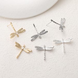 Op de afbeelding: Een verzameling oorbellen in de vorm van libellen, in goud- en zilverkleur. Elke oorbel heeft een gedetailleerd libellenontwerp met heldere edelsteenaccenten. De oorbellen worden gepresenteerd op een witte ondergrond.