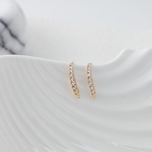 May include: A pair of gold-colored ear climbers, each adorned with a row of small, clear gemstones. The earrings are curved and sit on a white, textured surface, creating a contrast between the warm gold and the cool white background.
