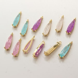May include: A collection of teardrop-shaped jewelry charms in various colors, including light blue, purple, pink, and clear. Each charm has a gold-colored metal frame and a small loop for attaching to a chain or bracelet. The charms are designed for jewelry making.