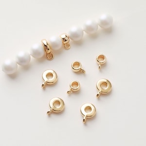 Pode incluir: Tampas de contas de metal dourado com um pequeno laço na parte superior. As tampas de contas estão dispostas sobre uma superfície branca com uma linha de pérolas brancas.