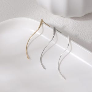 May include: Three pairs of earrings in gold and silver tones. The earrings have a long, curved design with a post backing. The earrings are displayed on a white, curved tray.