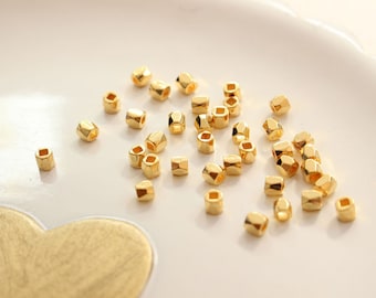 May include: A collection of gold-colored metal beads, each with a hexagonal shape. The beads are scattered on a white surface.