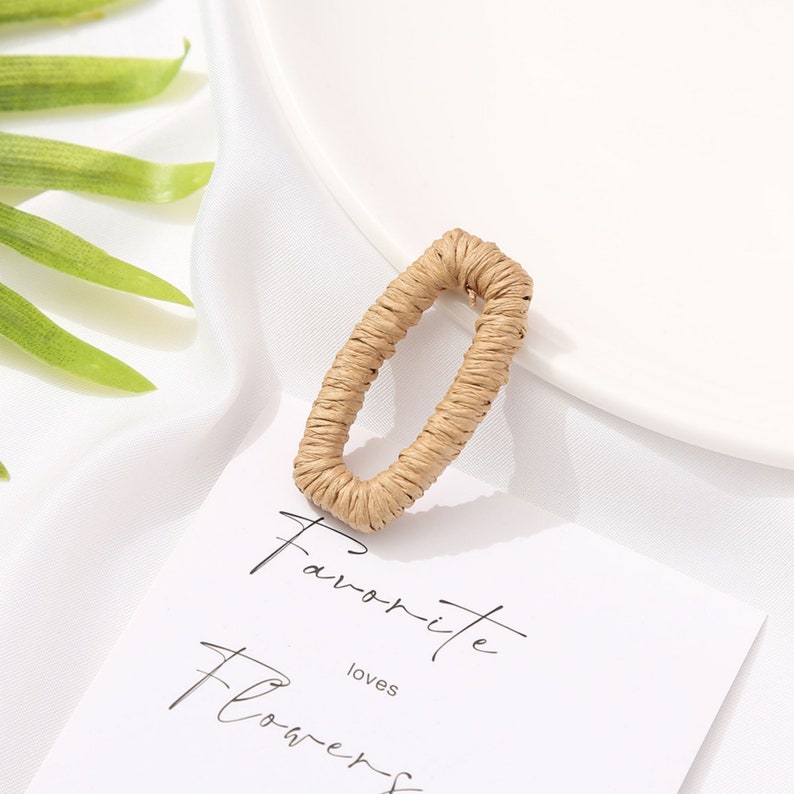 May include: A tan, woven, rectangular earring. The earring is made of a natural fiber and has a textured appearance. The earring is displayed on a white surface with a card that reads "Favorite loves Flowers".