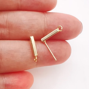 Pode incluir: Dois postes de brinco dourados com uma barra reta e um laço na ponta. Os postes são segurados na mão.