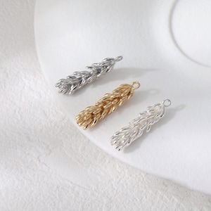May include: Three metal wheat charm pendants in silver and gold tones. The charms have a textured wheat stalk design. The pendants are displayed on a white surface.