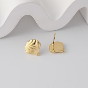 May include: Two gold-colored seashell stud earrings. The earrings have a detailed, textured design, with a post and a small loop for attaching to a chain or other jewelry. The earrings are on a white surface.