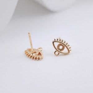 May include: A pair of gold-tone eye-shaped earring charms with a small loop at the top. The earrings are designed with a detailed outline of an eye with eyelashes.