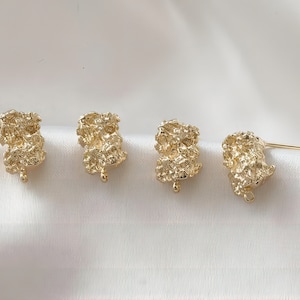 Puede incluir: Cuatro hallazgos de pendientes de color dorado con una superficie texturizada irregular. Los hallazgos tienen un bucle en la parte inferior y un poste en el lateral.