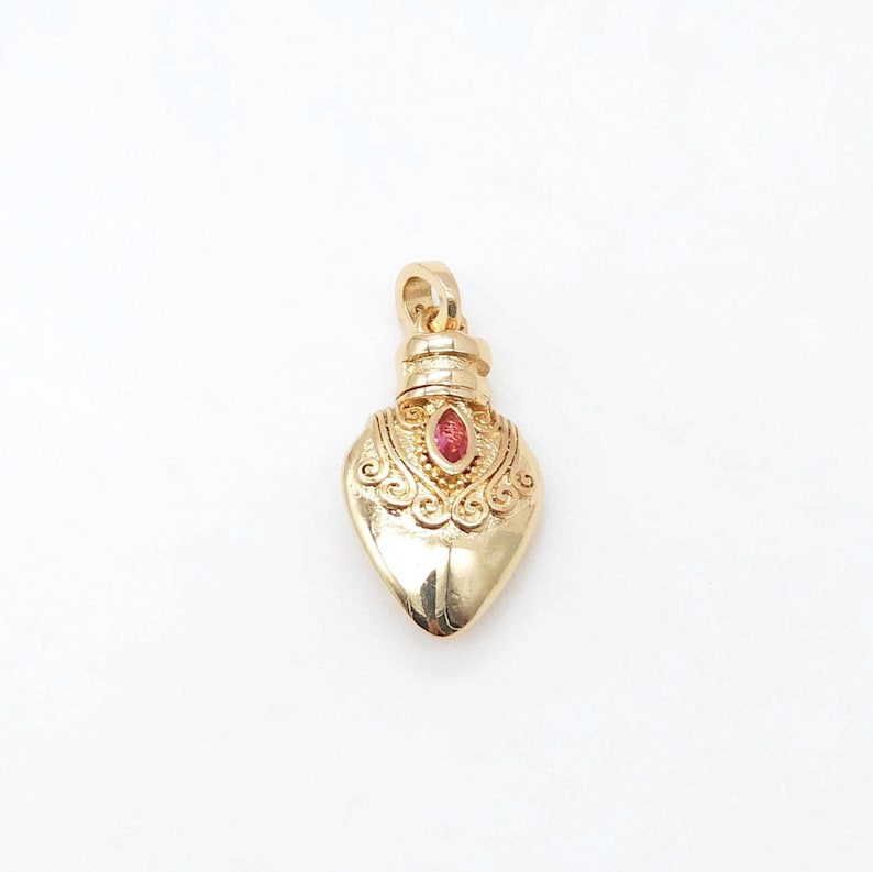 May include: A gold heart-shaped pendant with a red gemstone in the center. The pendant has intricate details and a loop at the top for attaching to a chain.