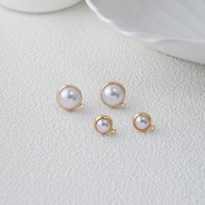 Peut inclure: Quatre boucles d'oreilles en perles dorées de différentes tailles sont disposées sur une surface blanche texturée. Chaque boucle d'oreille présente une perle blanche sertie dans un cadre doré, avec une petite boucle pour l'attache. Les perles ont un éclat brillant.