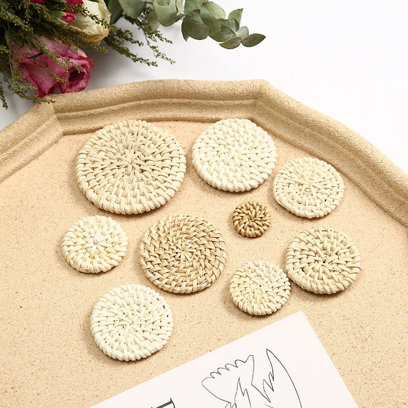 May include: A selection of woven rattan coasters in different sizes. The circular coasters have a natural beige hue and a textured surface. They are displayed on a light brown tray, with dried flowers visible in the background.