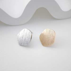 May include: A pair of small, geometric earrings. One earring is silver and the other is gold. Both earrings have a textured, brushed finish.