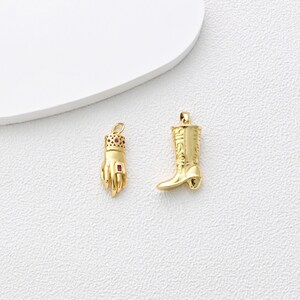 May include: Two gold-colored pendants. One is a hand wearing a crown and a ruby ring. The other is a cowboy boot with decorative detailing. Both pendants are on a white surface.