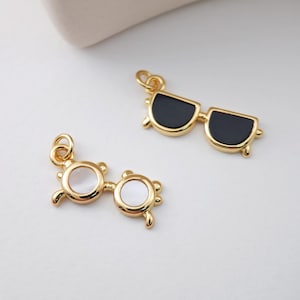 May include: Two gold-tone charm pendants in the shape of sunglasses. One pair has black lenses and the other pair has white pearlescent lenses.