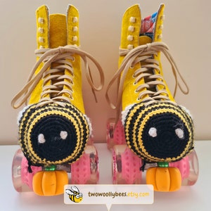 May include: A pair of yellow roller skates with tan laces and pink wheels. Each skate has a crocheted bee-themed toe stop cover with black and yellow stripes and white accents. Orange pumpkin-shaped toe stops are attached.