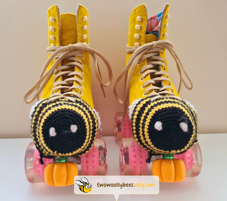 May include: A pair of yellow roller skates with tan laces and pink wheels. Each skate has a crocheted bee-themed toe stop cover with black and yellow stripes and white accents. Orange pumpkin-shaped toe stops are attached.