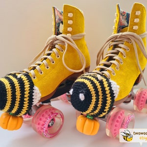 May include: A pair of yellow roller skates with tan laces and pink wheels. Each skate has a black and yellow crocheted bee-themed toe cap and an orange pumpkin-shaped toe stop. The interior lining of the skates features a colorful floral pattern.