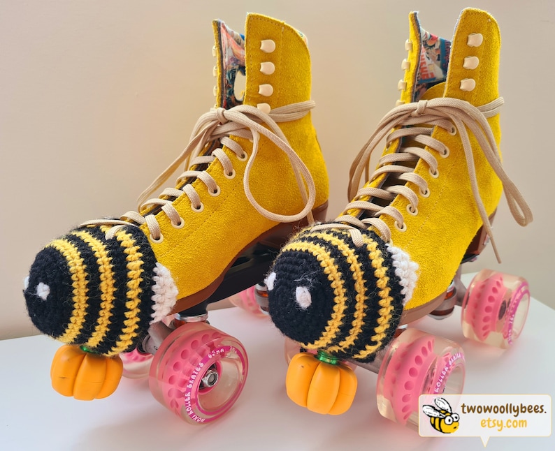 May include: A pair of yellow roller skates with tan laces and pink wheels. Each skate has a black and yellow crocheted bee-themed toe cap and an orange pumpkin-shaped toe stop. The interior lining of the skates features a colorful floral pattern.