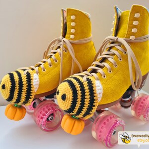May include: A pair of yellow roller skates with tan laces and black and yellow crocheted bee-themed toe caps. The wheels are pink and the toe stops are orange. The skates have a suede-like texture. The Etsy shop logo is in the bottom right corner.