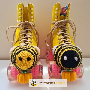 May include: A pair of yellow roller skates with tan laces and pink wheels. Each skate has a crocheted bee-shaped toe stop cover with black and yellow stripes. The toe stops are orange pumpkin-shaped.