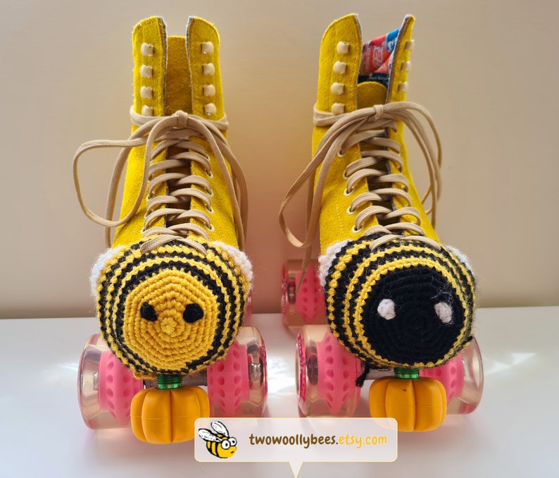 May include: A pair of yellow roller skates with tan laces and pink wheels. Each skate has a crocheted bee-shaped toe stop cover with black and yellow stripes. The toe stops are orange pumpkin-shaped.