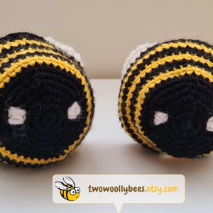 May include: Two handmade crochet bee toys. Each bee features black and yellow stripes, white accents, and white eye details. The toys are displayed on a white surface. The Etsy shop name, "two woolly bees", is displayed in a white box with a cartoon bee.