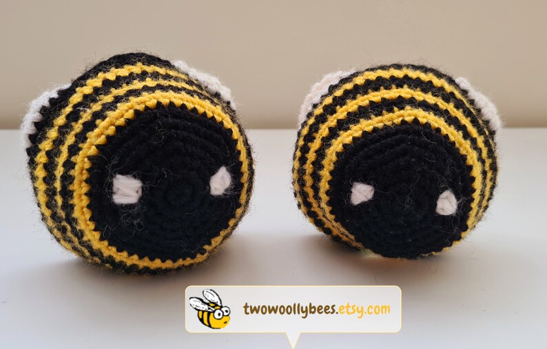 May include: Two handmade crochet bee toys. Each bee features black and yellow stripes, white accents, and white eye details. The toys are displayed on a white surface. The Etsy shop name, "two woolly bees", is displayed in a white box with a cartoon bee.