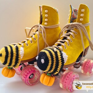 May include: A pair of yellow roller skates with tan laces and pink wheels. Each skate has a crocheted bee-themed toe stop cover with black and yellow stripes and a small orange pumpkin. The skates are decorated with a floral pattern.