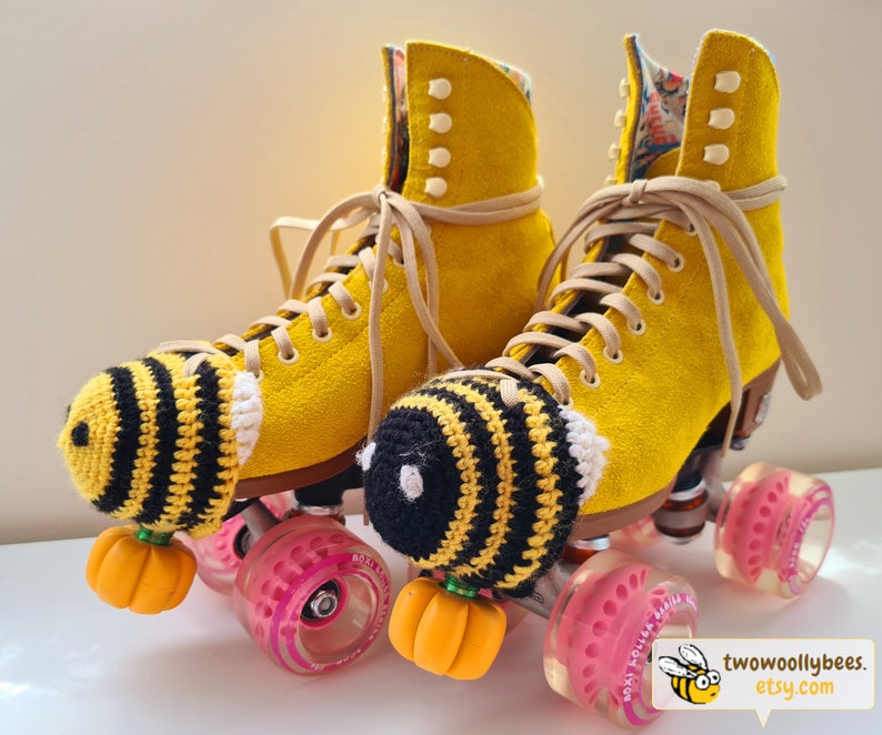 May include: A pair of yellow roller skates with tan laces and pink wheels. Each skate has a crocheted bee-themed toe stop cover with black and yellow stripes and a small orange pumpkin. The skates are decorated with a floral pattern.