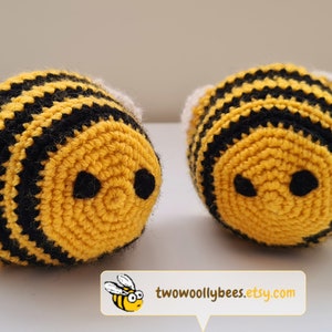 May include: Two handmade crochet bees. Each bee has a yellow body with black stripes, black eyes, and white wings. The bees are round and plush. The Etsy shop name, two woolly bees, is in the bottom right corner.