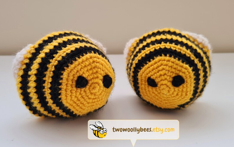 May include: Two handmade crochet bees. Each bee has a yellow body with black stripes, black eyes, and white wings. The bees are round and plush. The Etsy shop name, two woolly bees, is in the bottom right corner.