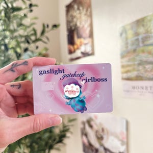 Peut inclure: Une carte rectangulaire avec les mots "gaslight gatekeep girlboss" dans un design rose et violet. Une créature de dessin animé est au centre de la carte. La carte est tenue dans une main.