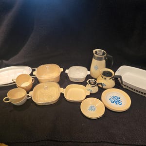 May include: A collection of vintage kitchenware in shades of yellow and white, including casserole dishes, cups, plates, a teapot, and a pitcher. The pieces feature a blue floral design. The items are arranged on a dark surface.