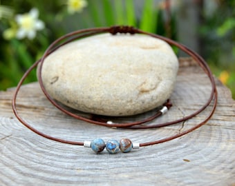 Può includere: Una collana in pelle marrone con tre perline blu e marroni. La collana è esposta su una pietra chiara, con una superficie in legno e vegetazione sullo sfondo.