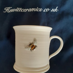 May include: Cream-coloured ceramic mug featuring a bumblebee motif. The mug has a curved handle and a flared rim. The bumblebee is detailed with brown and yellow stripes and white wings. The text "Hewittceramics.co.uk" is visible in the background.