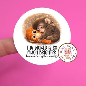 May include: A circular sticker with a sleeping brown monkey embracing an orange plush toy. The text "THE WORLD IS SO MUCH BRIGHTER because you exist" is below the image. A smaller logo says "THE GIRL BOSS CAFE".