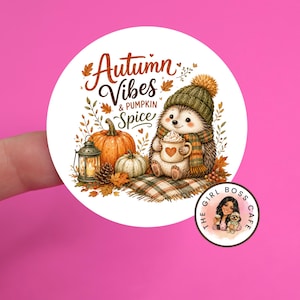 May include: A round sticker featuring a hedgehog wearing a green knit hat and scarf, holding a mug with a heart. The text reads Autumn Vibes & Pumpkin Spice. It includes a pumpkin, a lantern, and autumn leaves on a plaid blanket. A small logo is present.