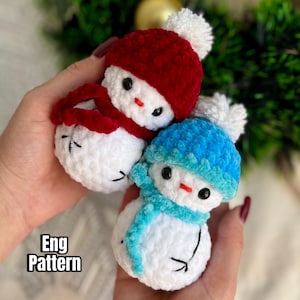 Puede incluir: Dos adornos de muñeco de nieve de crochet, uno con un gorro y una bufanda rojos y el otro con un gorro y una bufanda azules. Los muñecos de nieve son blancos con ojos negros y una nariz roja. El texto "Eng Pattern" es visible en la imagen.