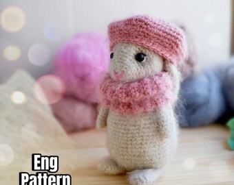 Może przedstawiać: Pluszowa zabawka amigurumi w kształcie myszy, ubrana w różowy kapelusz i szalik. Zabawka leży na drewnianej powierzchni.