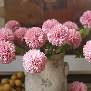 May include: A bouquet of pink artificial flowers in a grey ceramic vase. The flowers are arranged in a cluster and have a fluffy, pom-pom like texture.