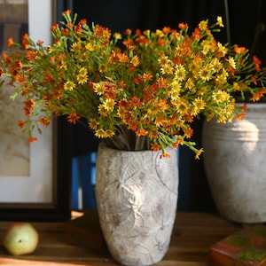 Puede incluir: Un ramo de flores artificiales en un jarrón. Las flores son una mezcla de amarillo y naranja, y tienen un aspecto delicado y vaporoso.