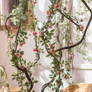 Puede incluir: Enredadera artificial de rosas con flores rosas y hojas verdes envueltas alrededor de una rama marrón. La enredadera cuelga del techo y es un elemento decorativo para un hogar o evento.