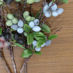 May include: A bunch of artificial green and blue berries on brown twigs with green leaves. The berries are clustered together and look realistic.