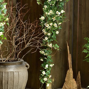 Puede incluir: Una vid artificial de flores blancas con hojas verdes colgando de una pared de madera. La vid est&aacute; colgada de un gancho y est&aacute; rodeada de piedras blancas y una maceta marr&oacute;n.